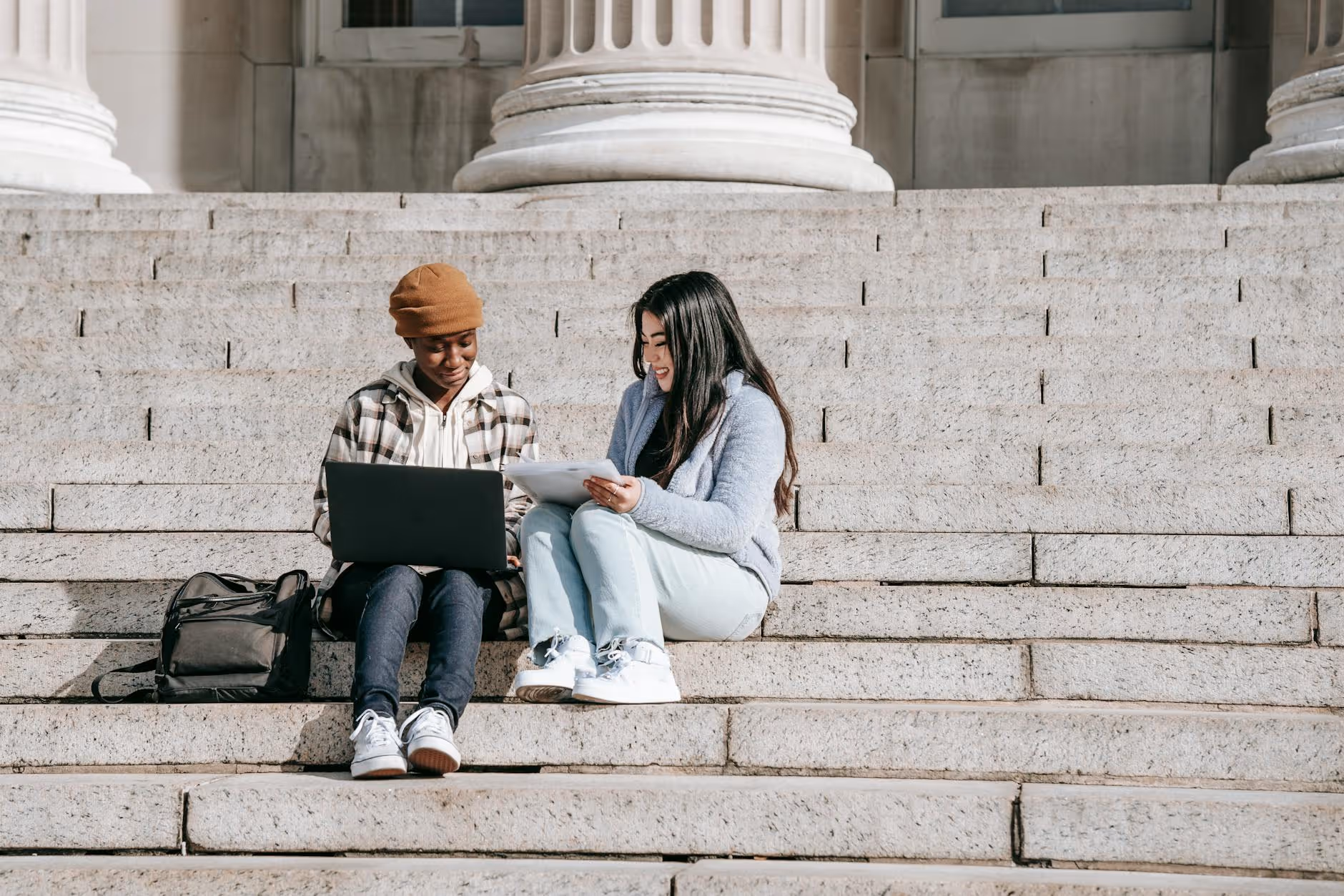 2 people sitting together on stairs working on laptop