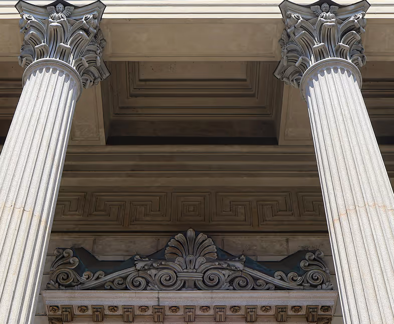 A symmetrical, low-angle close-up of classical architecture, featuring two fluted columns with highly ornate Corinthian capitals supporting a stone entablature. Below the columns, there is elaborate relief carving over a doorway, including a central shell or fan motif framed by stylized scrolls and a band of Greek key (meander) pattern above it. The stonework is a light tan or beige color.