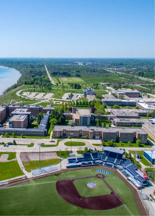 An aerial view of a large, flat college campus on a clear, sunny day. In the foreground is a baseball stadium with an infield of brown dirt and green outfield grass, complete with blue seating stands. The middle ground features several institutional buildings with brown roofs, interspersed with green lawns and trees. A wide river runs along the left side of the campus, and the surrounding area is characterized by flat, wooded terrain extending to the horizon.