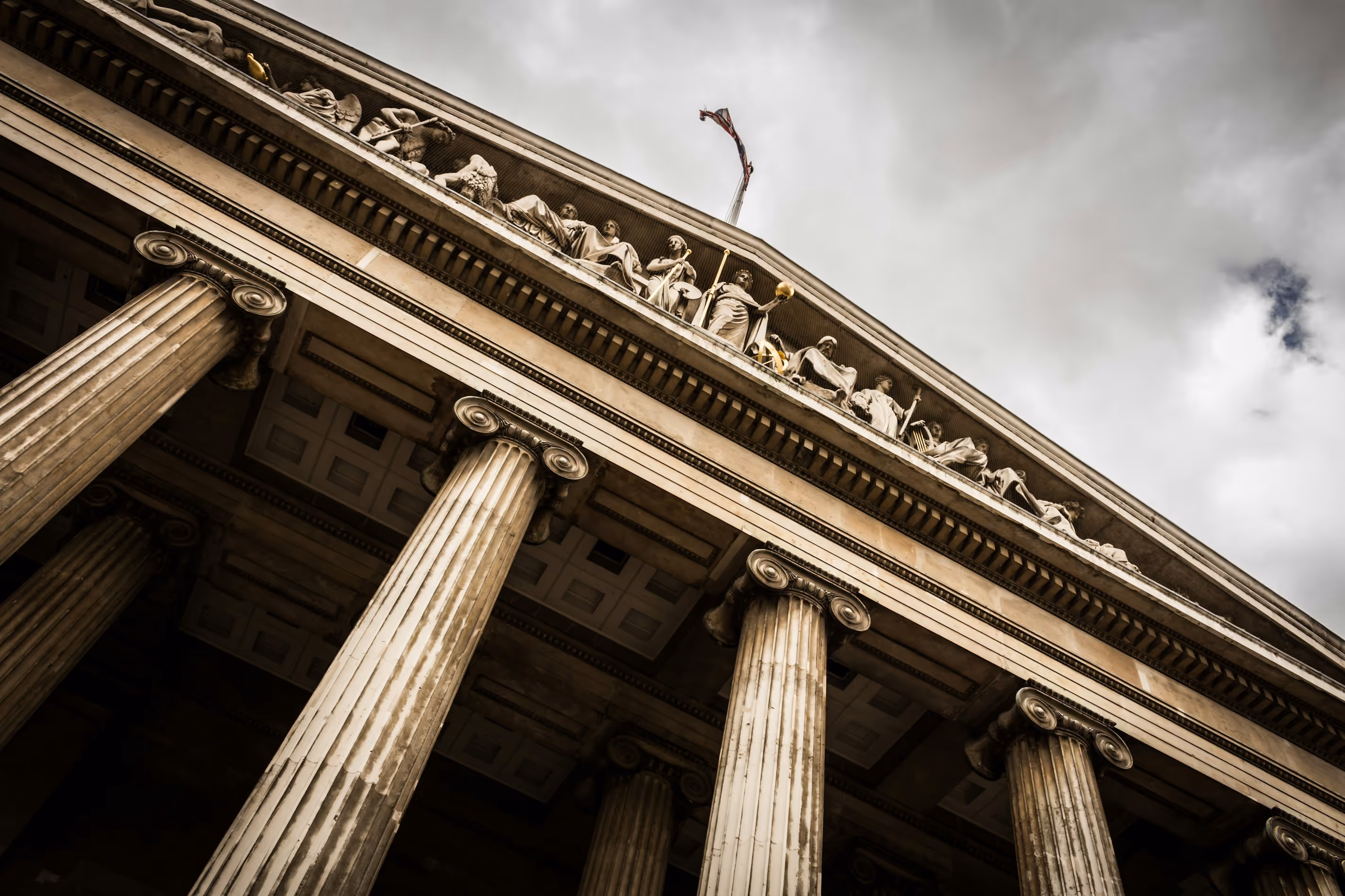 Low-angle view of ornate classical columns supporting a building facade with sculpted figures and a flag above.